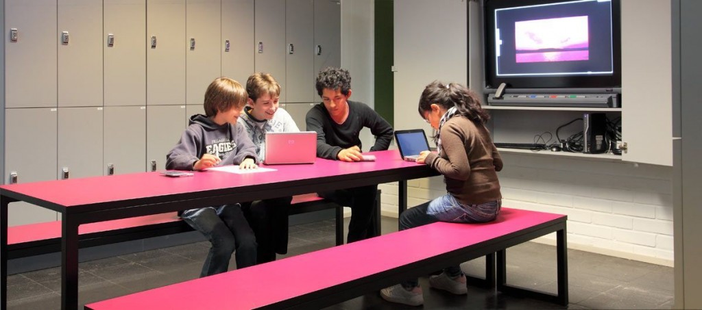 Children are sitting in a hall, working on computers. The benches are red and we can see another big screen in the background. 